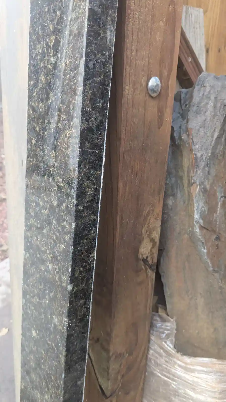 A close-up photo shows a dark, speckled granite slab standing next to a rough, brown wooden plank with a metal rivet. The…