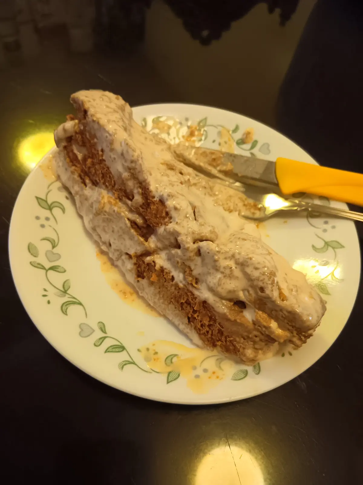 A slice of cake with light brown frosting sits on a white plate with green floral patterns, partially eaten with a knife and…