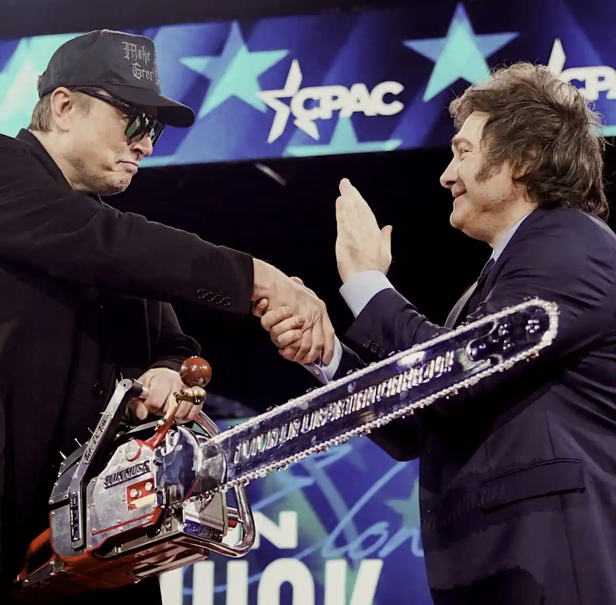 Two men are shaking hands on a stage with a CPAC logo in the background. The man on the left, wearing a black cap and…