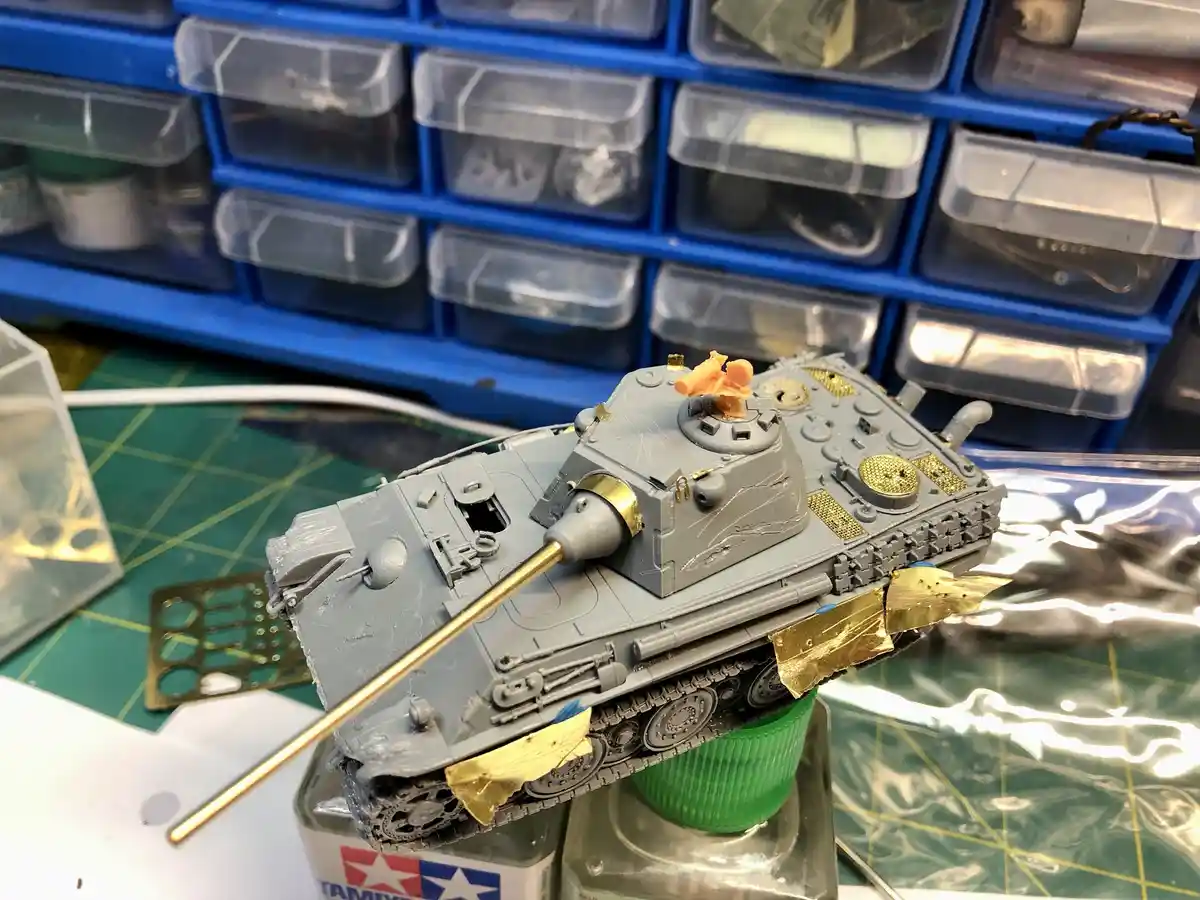 A grey scale model tank, likely a Tiger I, is shown on a workbench with blue storage drawers in the background. The tank is…