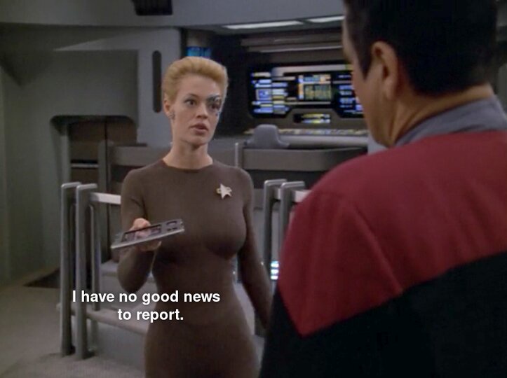 A woman in a brown Starfleet uniform with a star pin holds a PADD and looks concerned while facing a man in a red and black…