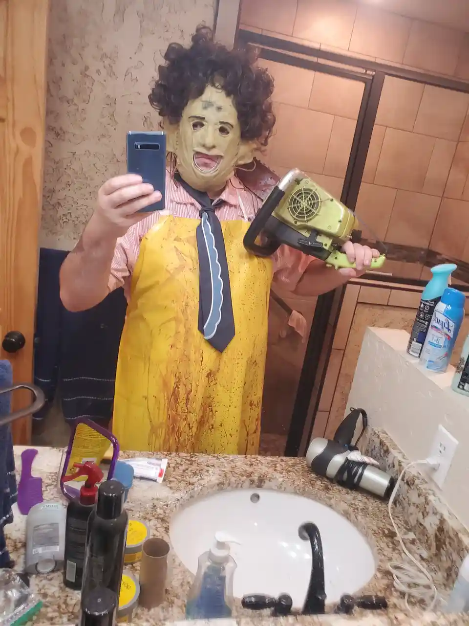 A person in a bathroom mirror selfie wearing a yellow apron splattered with fake blood, a grotesque mask with curly hair…