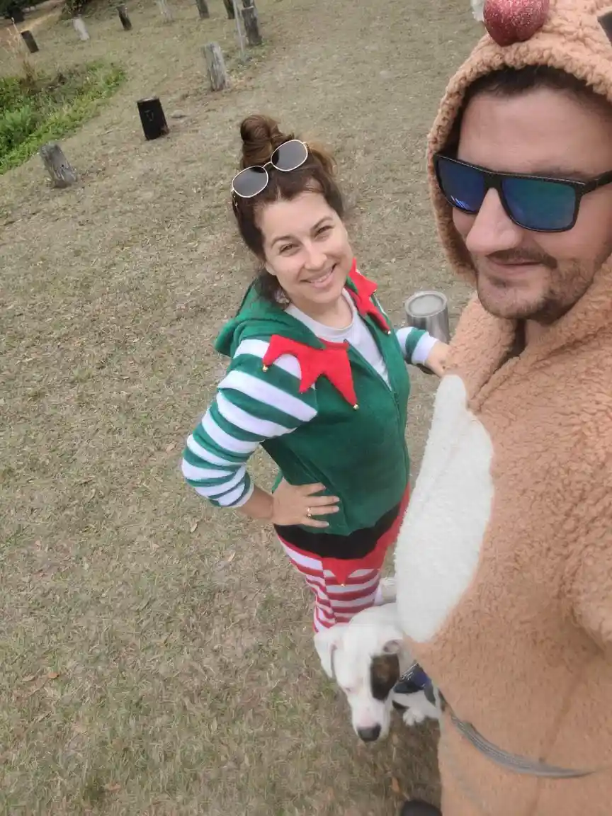 A man in a brown reindeer onesie with a red nose and sunglasses takes a selfie with a woman in a green elf costume with…