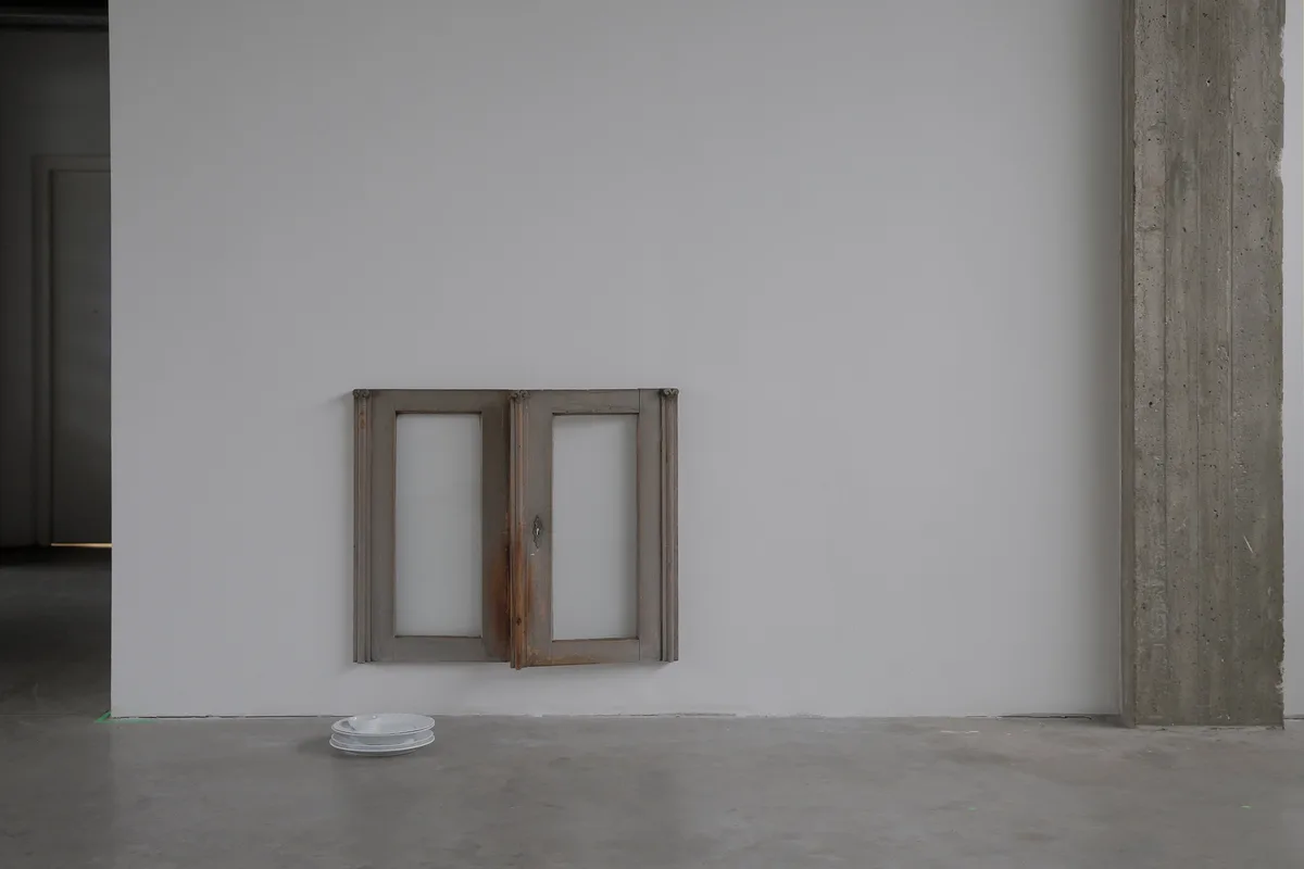 A minimalist art installation shows a small, weathered wooden window frame mounted on a plain white wall, with a stack of…