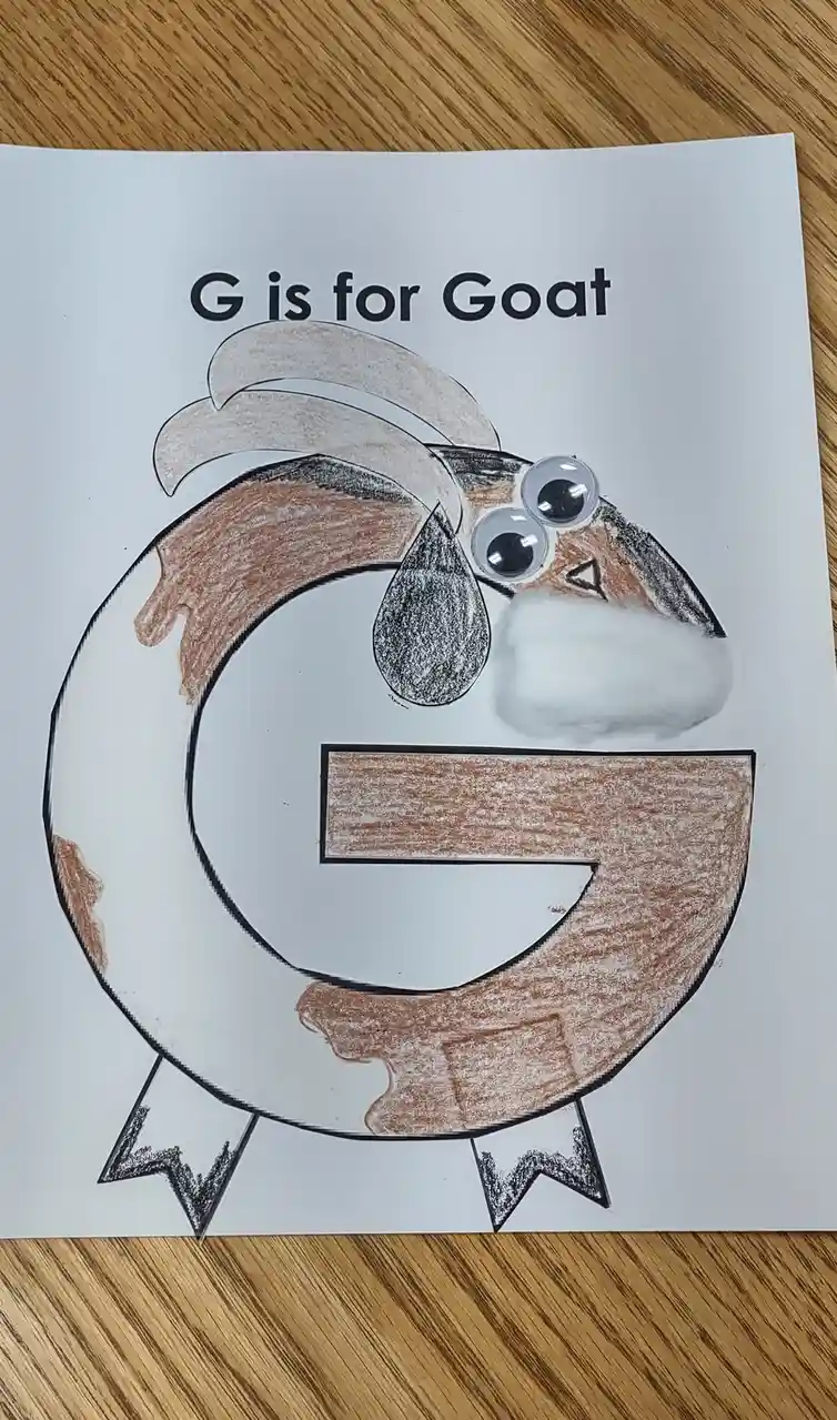 A child's craft project shows a large, brown-colored letter 'G' drawn on white paper, decorated with googly eyes, a cotton…