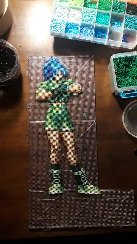 A handmade perler bead artwork of an anime-style character with blue hair and green outfit, created on a transparent…
