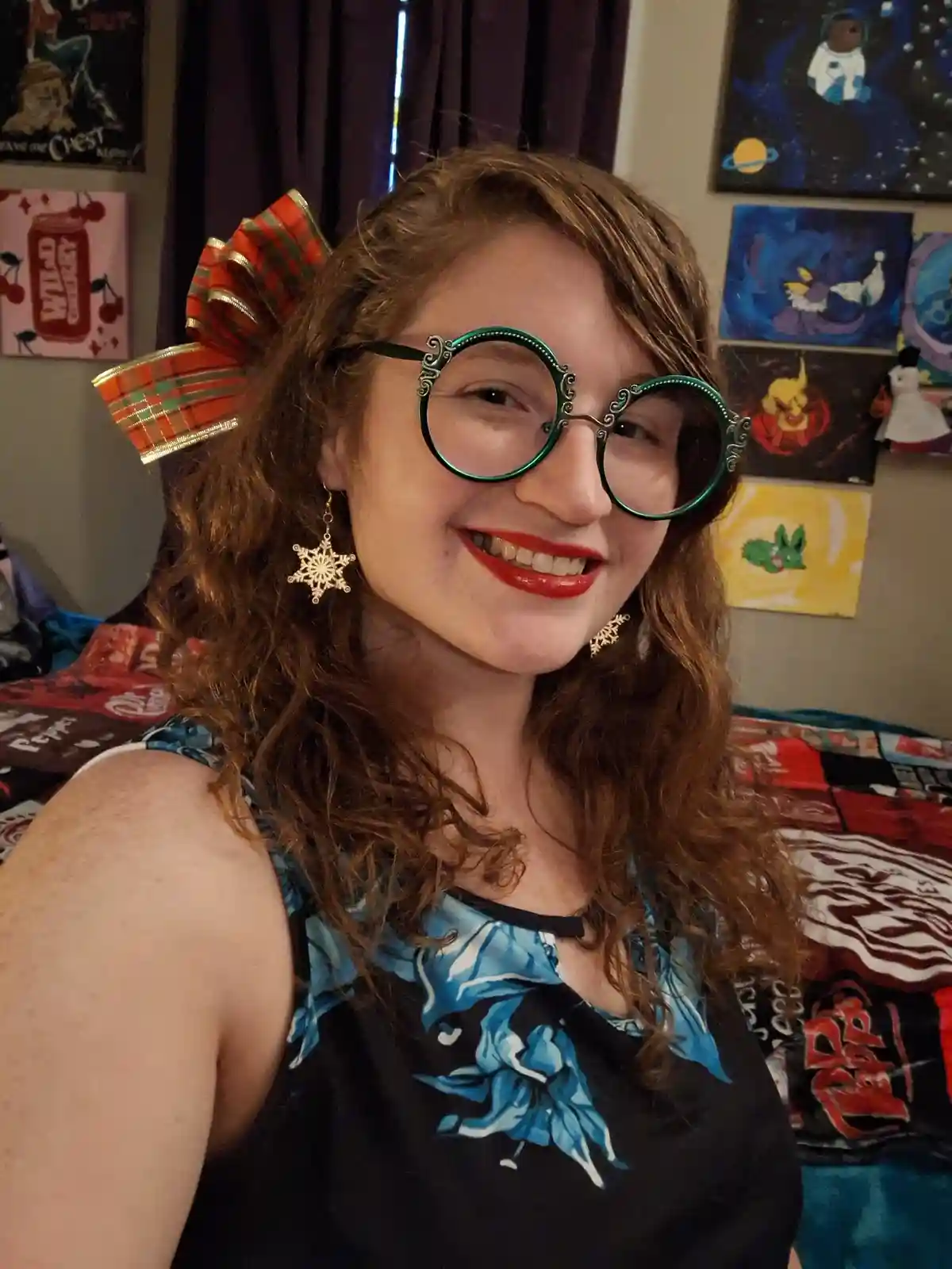 A young woman with curly brown hair smiles at the camera while wearing large, ornate green-rimmed glasses and snowflake…