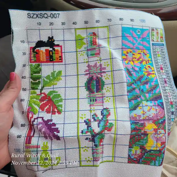 A hand holds up a partially completed cross-stitch pattern on white fabric, showing colorful stitched sections including a…