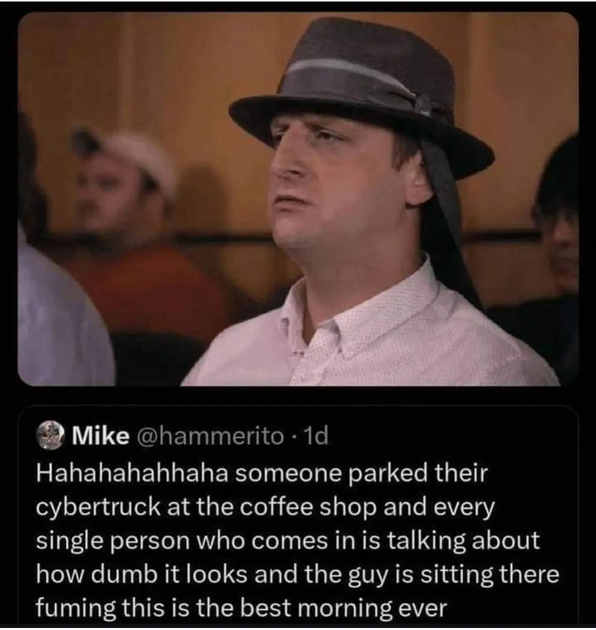 A man wearing a grey fedora and light pink shirt looks annoyed while sitting in a coffee shop, with blurred people in the…