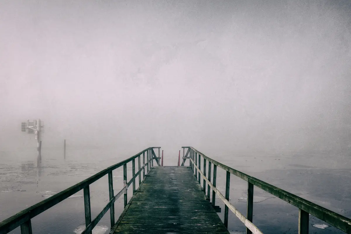 A foggy, atmospheric photo shows a wooden pier extending into a mist-covered body of water, with the path disappearing into…