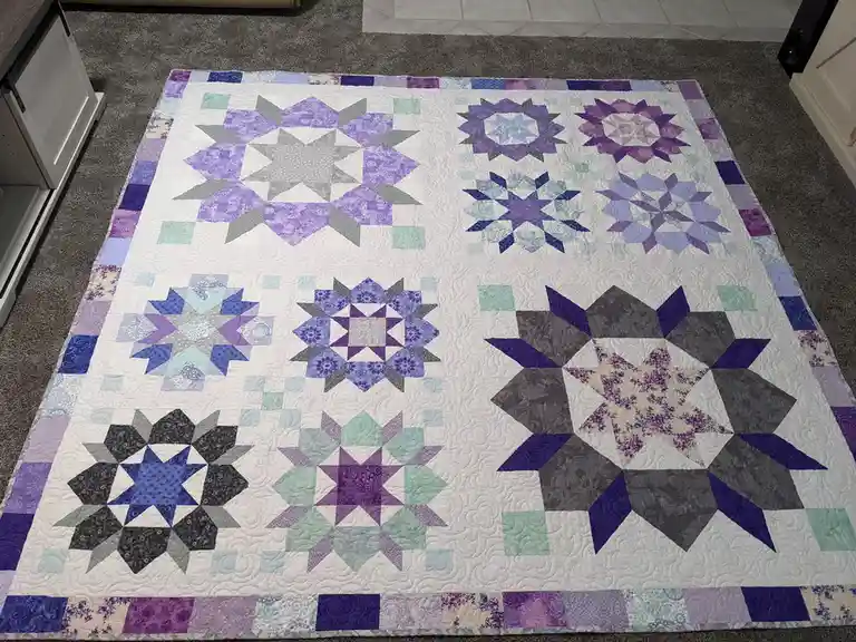 A large, handmade quilt with multiple star and floral patterns in shades of purple, blue, and gray is spread out on a…