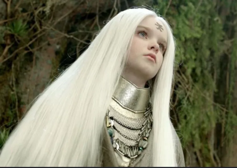 A woman with long white hair and a silver collar looks upward with a skeptical expression, her forehead marked with a…
