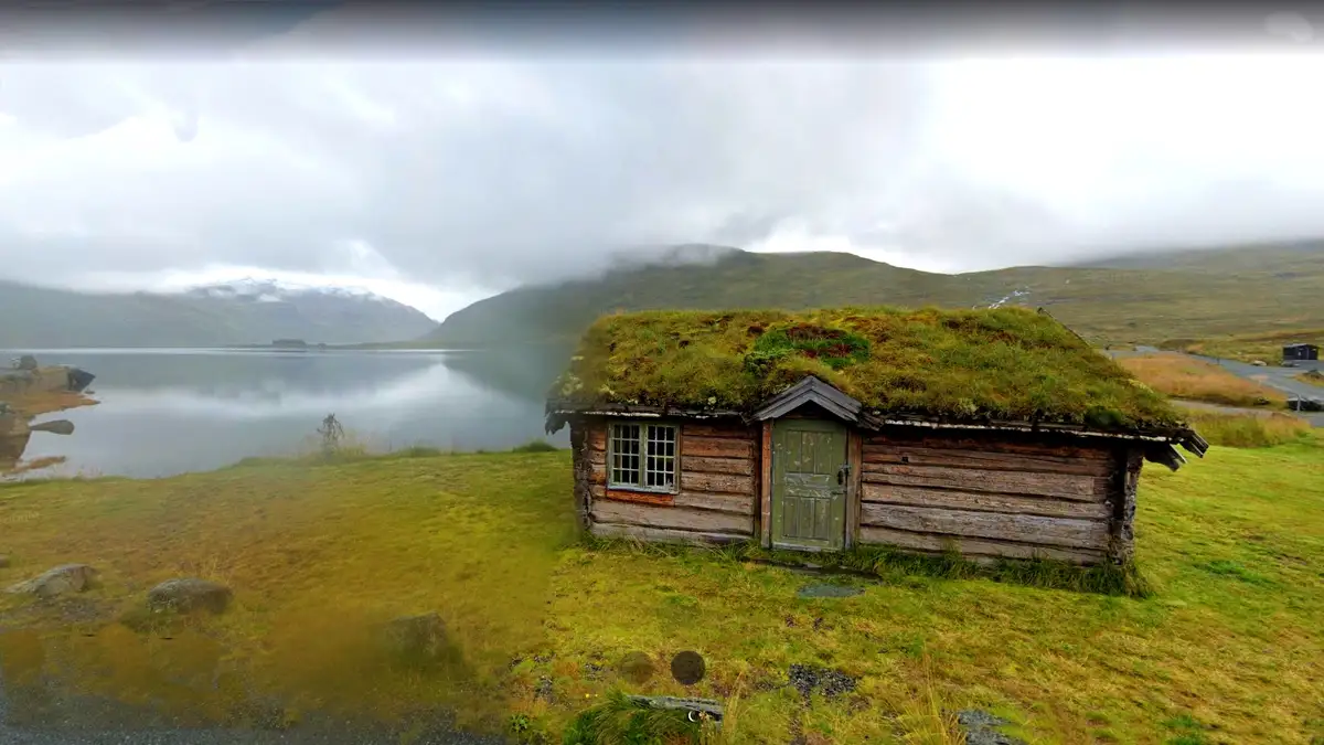 A rustic wooden cabin with a grassy roof sits beside a calm lake, surrounded by misty mountains under an overcast sky. The…