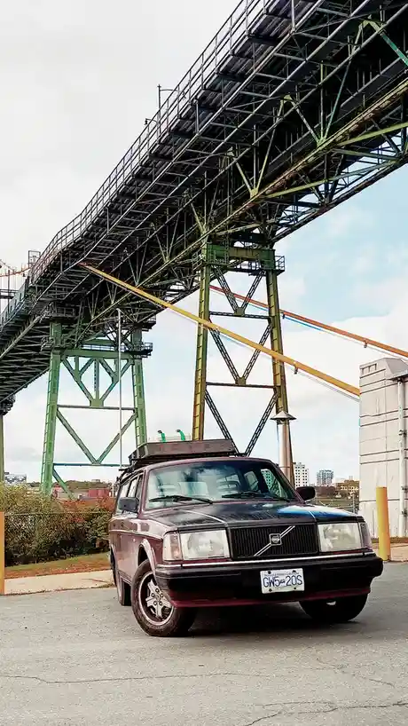 A maroon Volvo station wagon with a roof rack is parked under a large green steel bridge. The car has a British Columbia…
