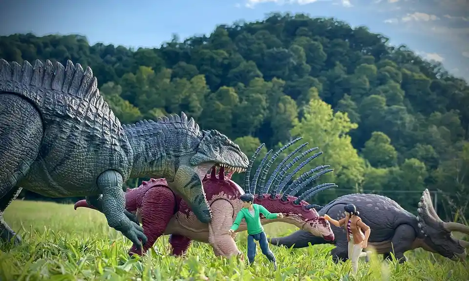A photo of toy dinosaurs and human action figures staged in a grassy field with trees in the background. A large grey…