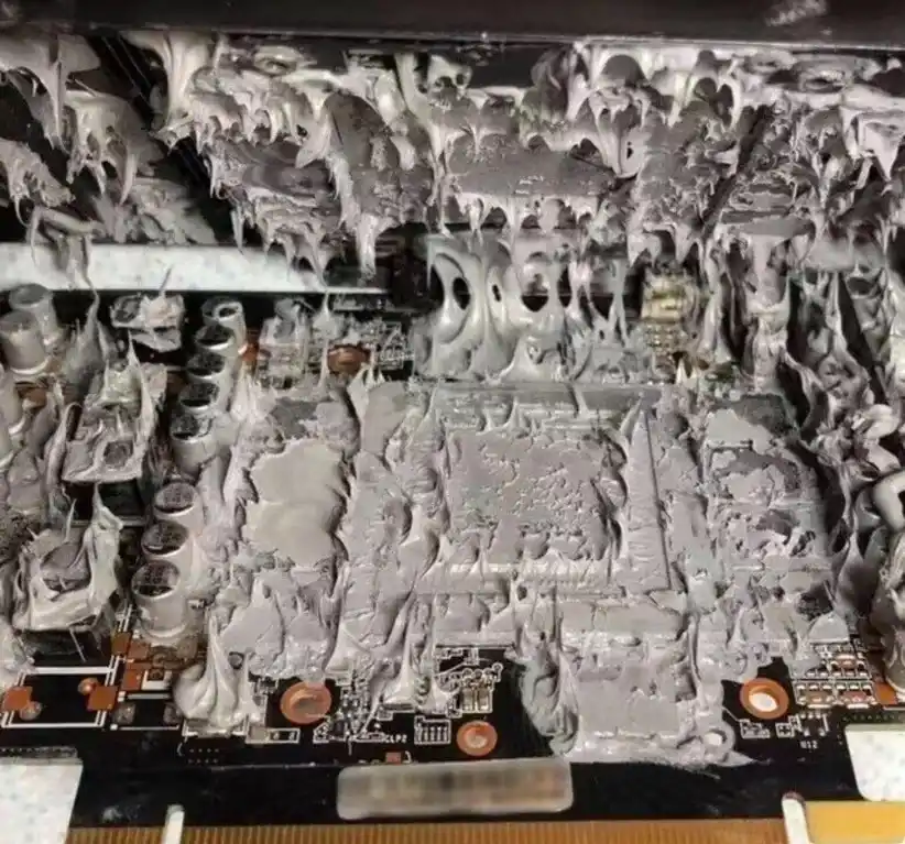 a close-up photograph shows a computer motherboard entirely engulfed in thick, gray thermal paste that has dripped and…