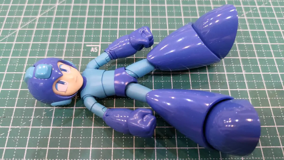 A blue and light blue Mega Man action figure lies on its back on a green cutting mat with grid lines, arms bent at the…