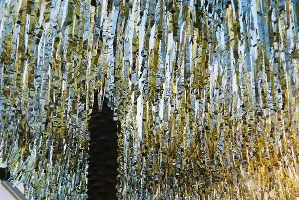 A close-up of shimmering gold and silver metallic streamers hanging vertically, with a dark, textured vertical object…