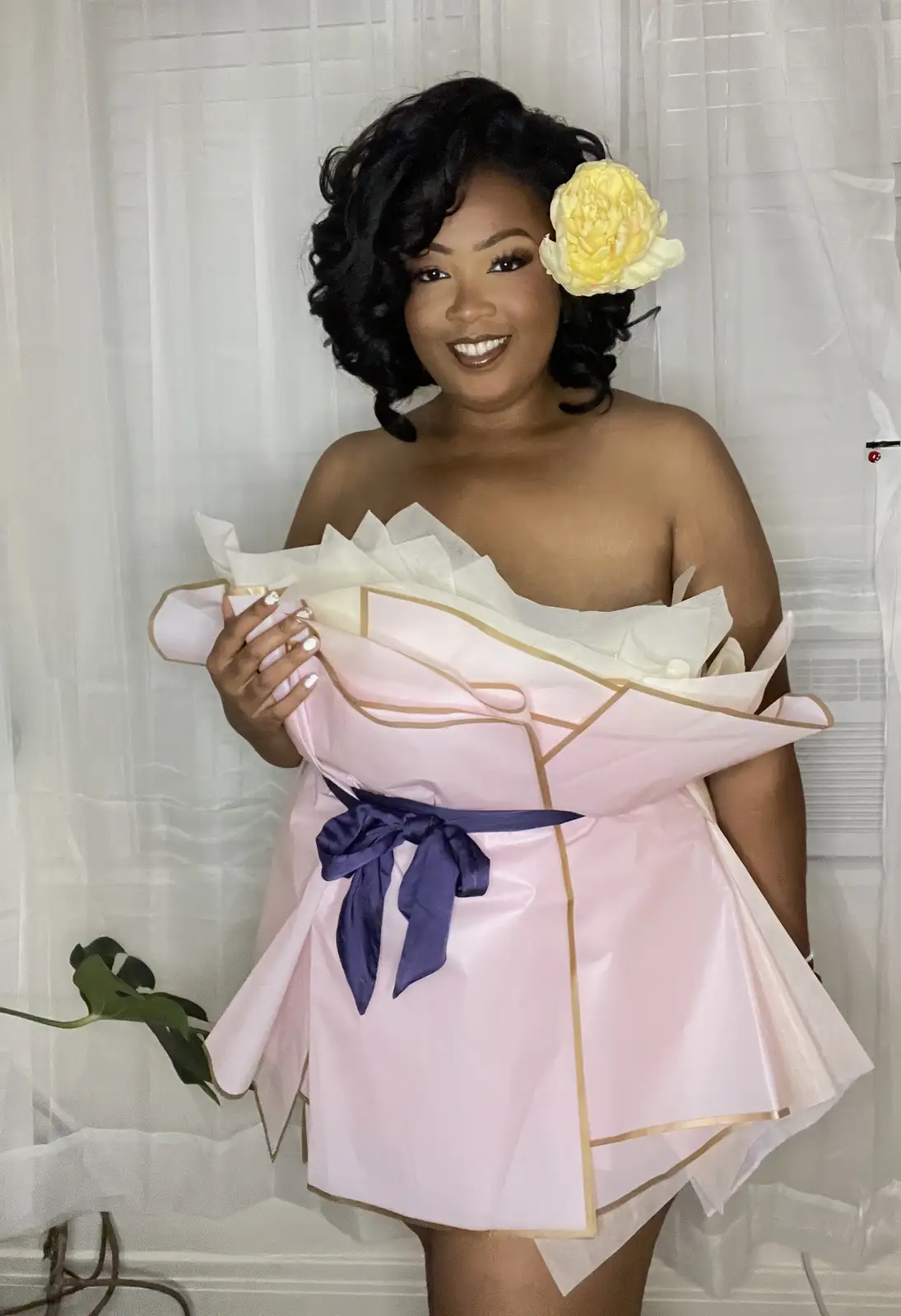 A smiling woman with a yellow flower in her hair poses while wearing a pink, bouquet-style dress made of wrapping paper with…