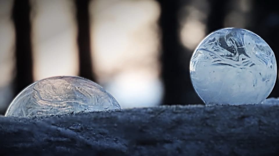 Two ice spheres sit on a snowy surface with a blurred forest background. The larger, more complete sphere on the right is…