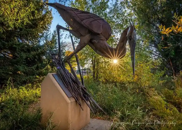 A large, dark metal sculpture of a heron stands on a concrete pedestal in a wooded area, with the sun shining brightly…