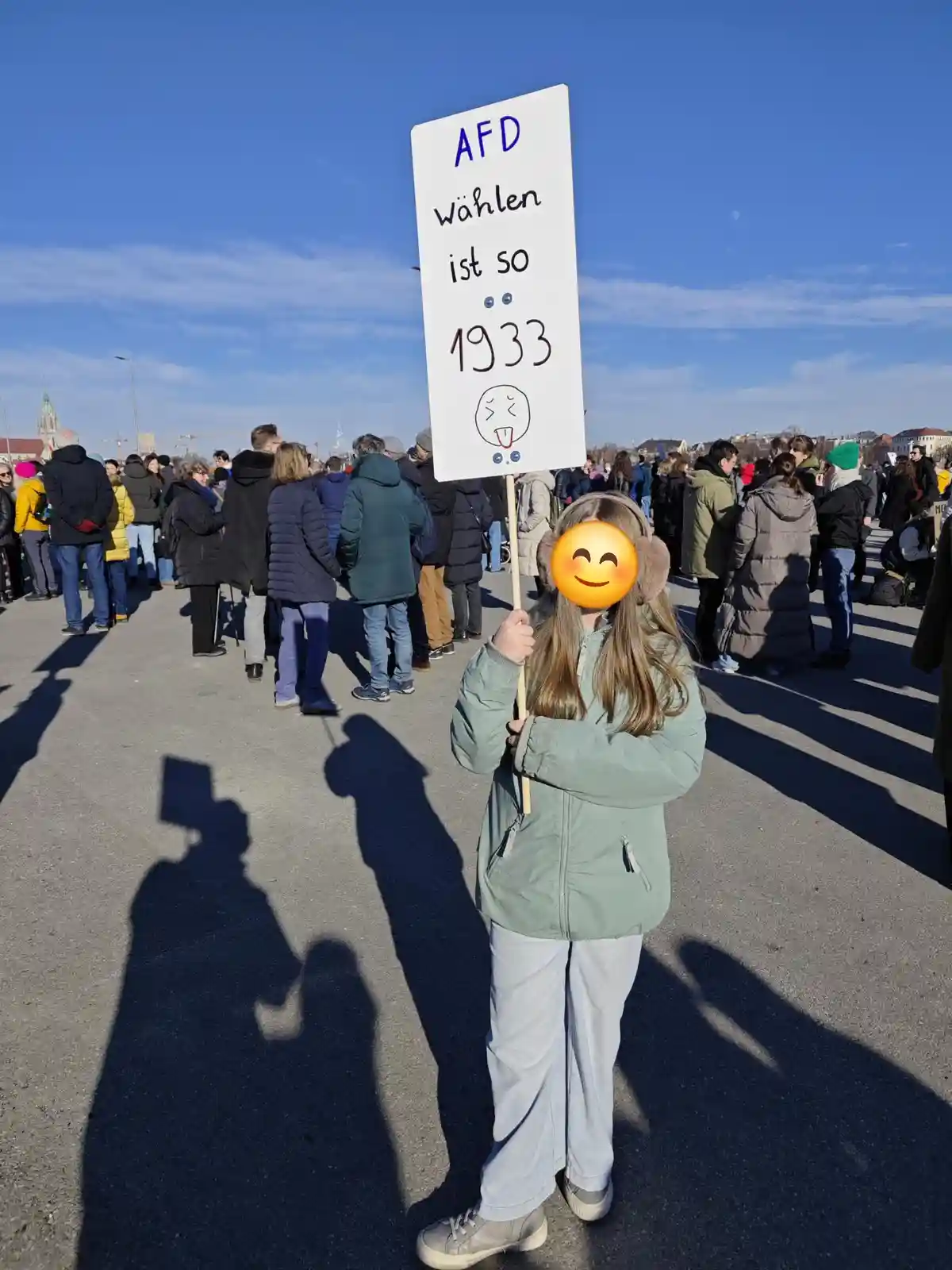 A young girl in a green jacket and earmuffs holds a handmade protest sign at an outdoor gathering under a clear blue sky…