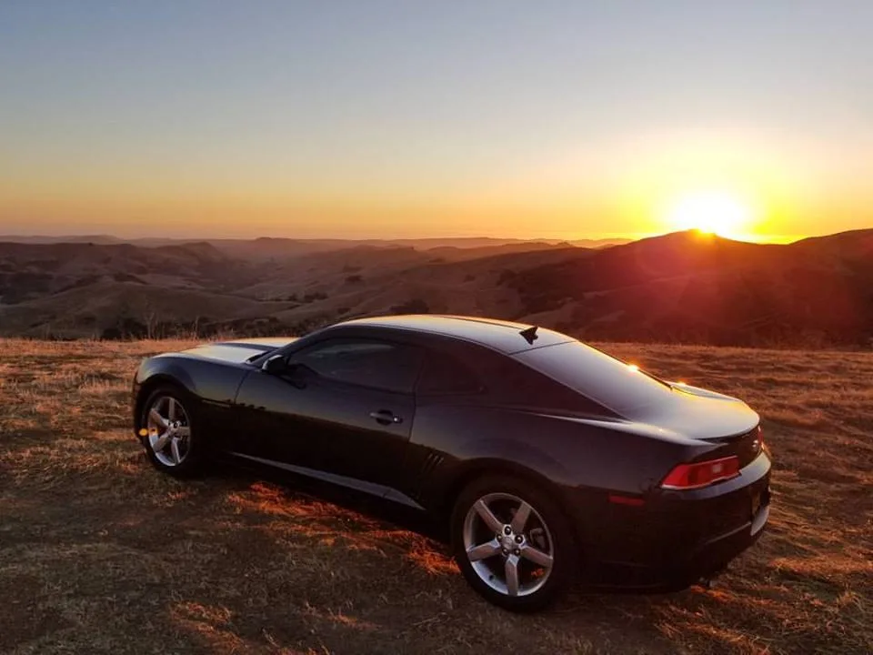 A black Chevrolet Camaro is parked on a grassy hilltop overlooking rolling hills at sunset, with the sun low on the horizon…