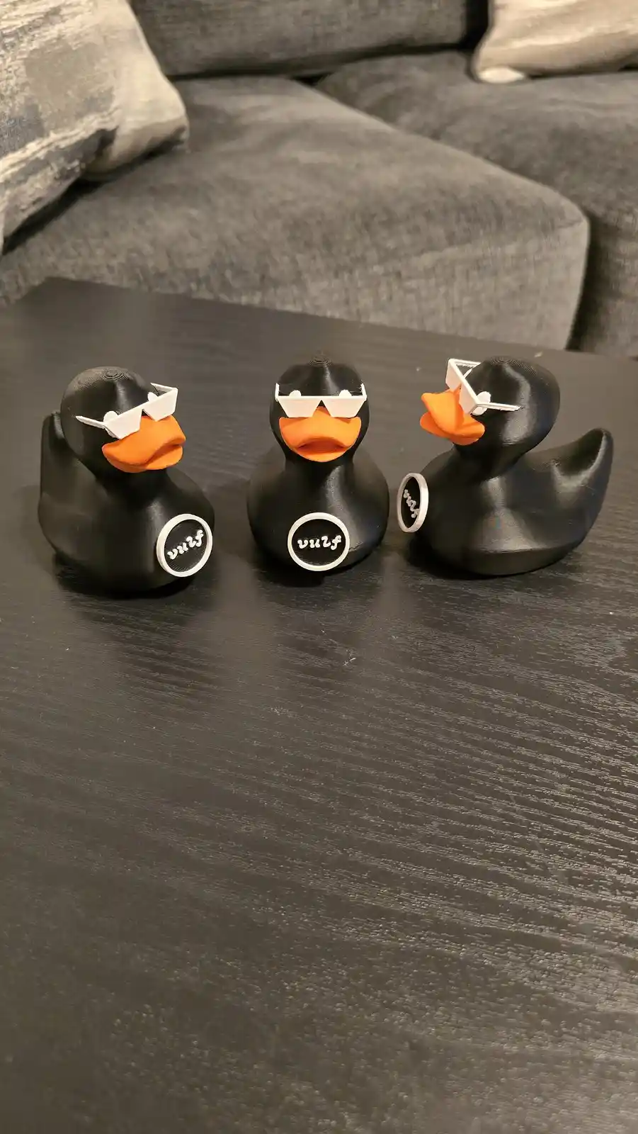 Three 3d-printed black rubber ducks with orange beaks sit on a dark wooden table, each wearing white 3d-printed sunglasses…