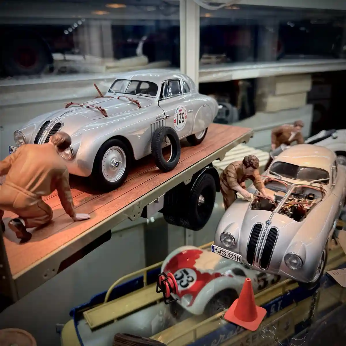 A photo of detailed scale model cars and figurines in a display case, showing miniature mechanics working on vintage racing…