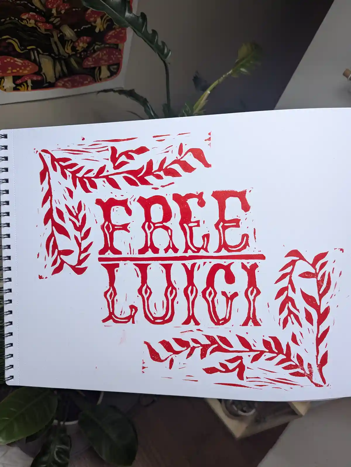 A red ink print on white paper in a spiral-bound sketchbook shows the words 'FREE LUCY' in a stylized, blocky font, framed…