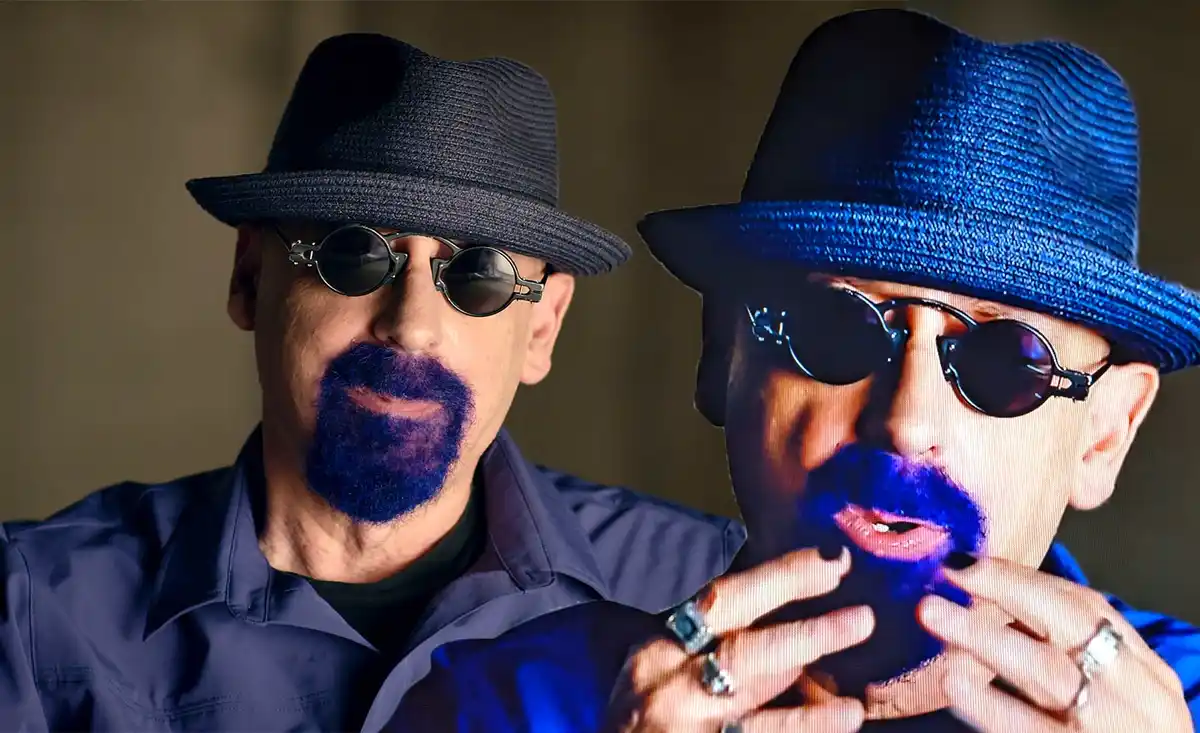 A man with a dark fedora and round sunglasses has a blue beard and is looking at a screen showing a magnified version of…