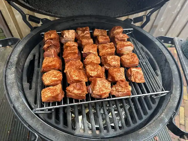 A top-down view of a black charcoal grill with its lid open, showing numerous seasoned meat cubes arranged neatly on the…