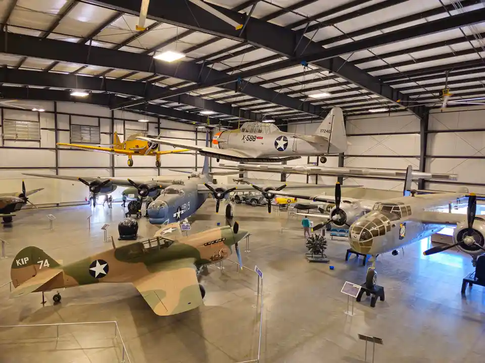 A wide shot of a large, well-lit aircraft museum hangar filled with numerous vintage military planes, some suspended from…