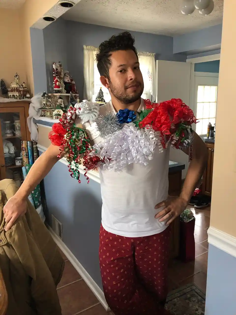 A man in a white t-shirt and red patterned pajama pants stands in a home decorated for Christmas, with festive ornaments and…