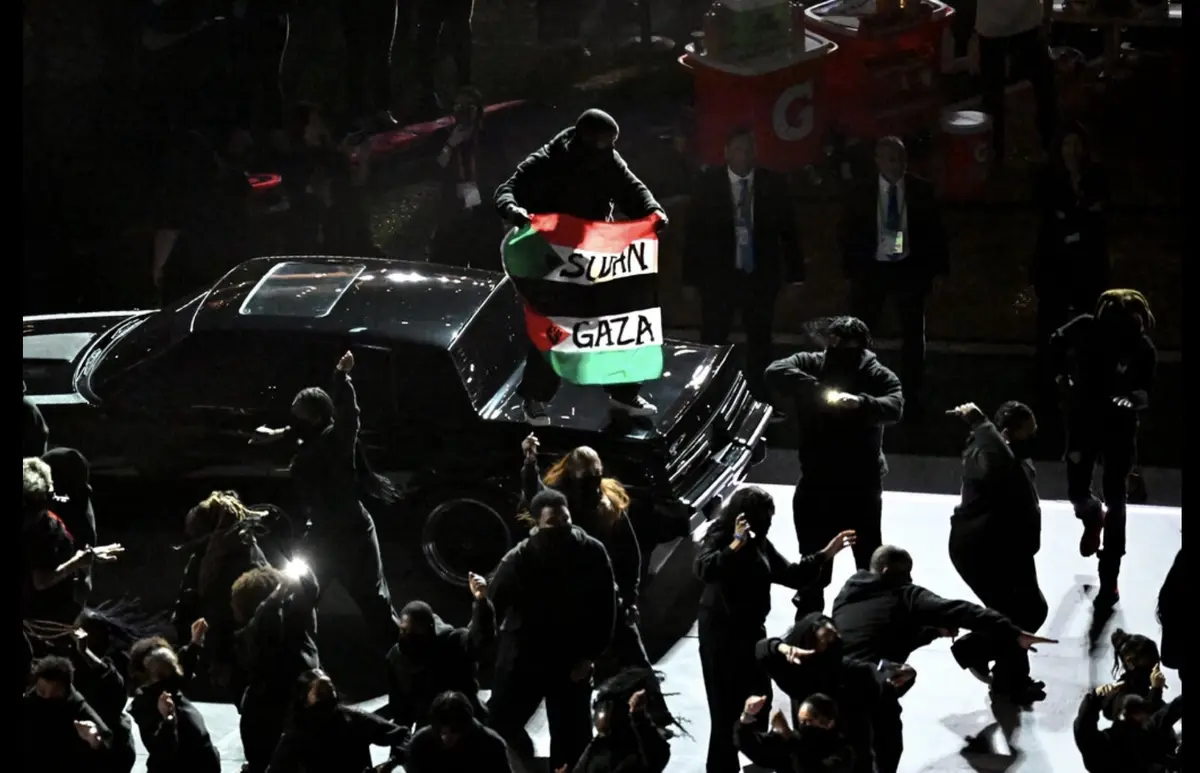 A person stands atop a black car on a stage, holding a Palestinian flag with the words 'SUDAN' and 'GAZA' written on its…