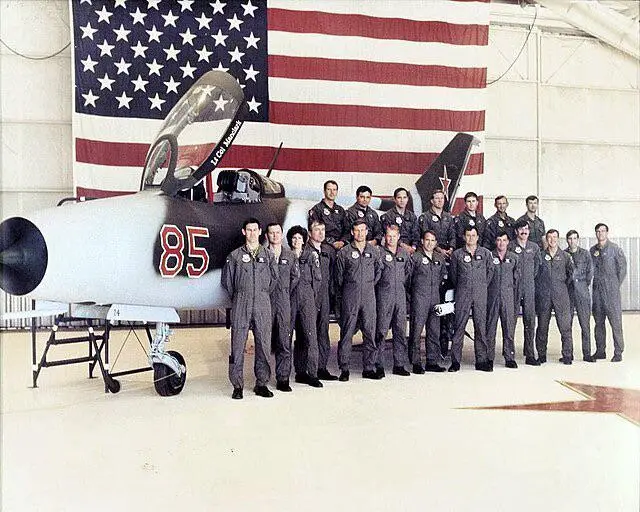 A vintage group photo shows a squadron of military pilots in uniform posing in front of a fighter jet marked with the number…