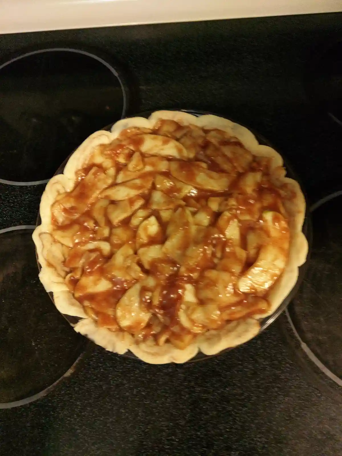 An uncooked apple pie with a fluted crust sits on a dark stovetop, filled with sliced apples coated in a glossy brown sugar…