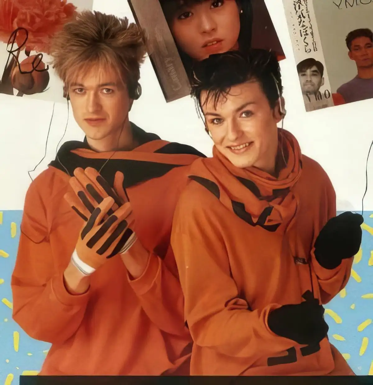 Two young men in matching orange hoodies with black accents and fingerless gloves pose against a wall covered in posters…
