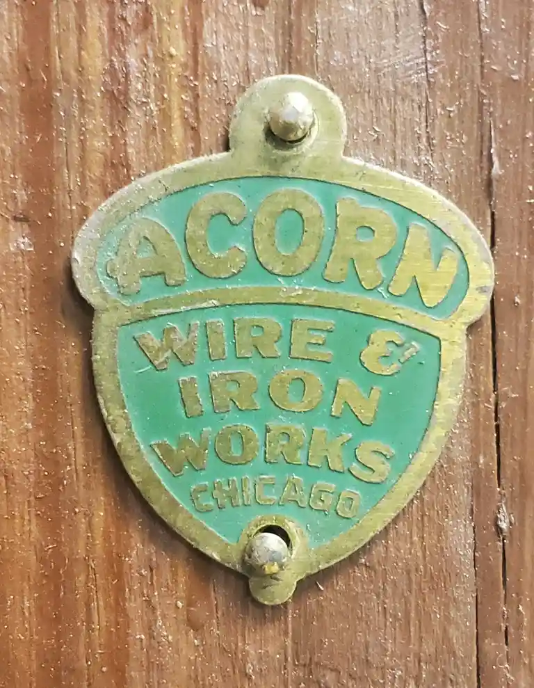 A close-up of a weathered, green-painted metal plaque shaped like an acorn, mounted on a wooden surface. The plaque reads…