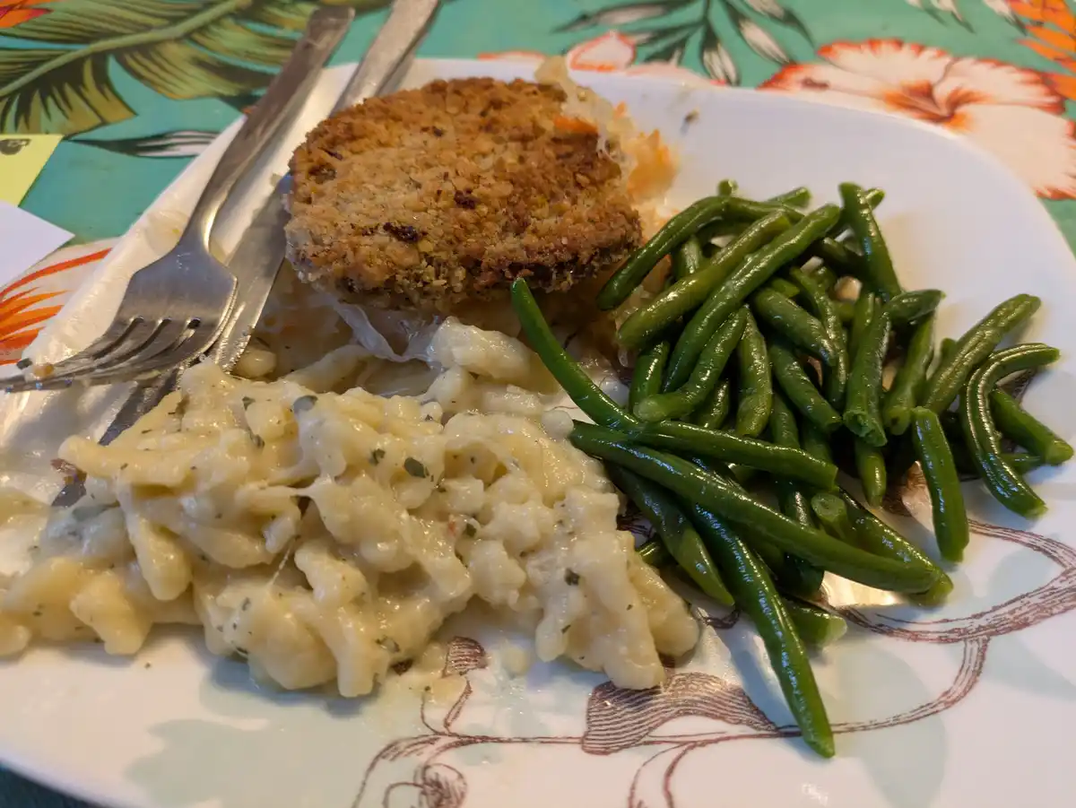 A plate of food featuring a breaded patty, green beans, and macaroni and cheese, with a fork and knife beside it. The…