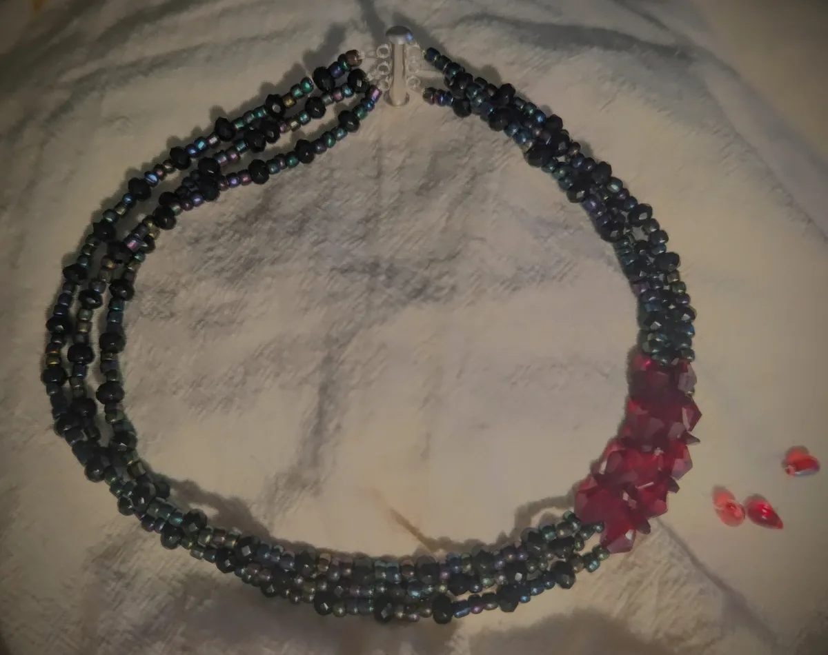 A handmade multi-strand necklace made of iridescent dark beads with a cluster of red crystal beads at one end, lying on a…