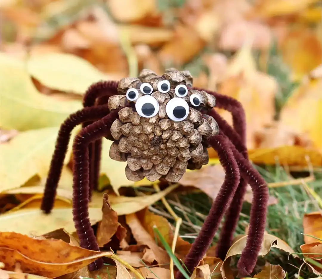 A handmade craft spider made from a pinecone body with multiple googly eyes and dark purple pipe cleaner legs, sitting among…