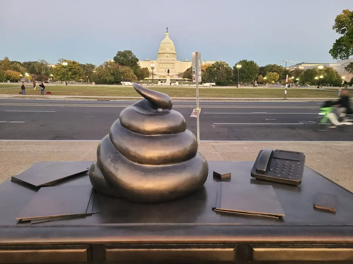A bronze sculpture of a poop emoji sits on a desk with a phone and papers, with the U.S. Capitol building visible in the…
