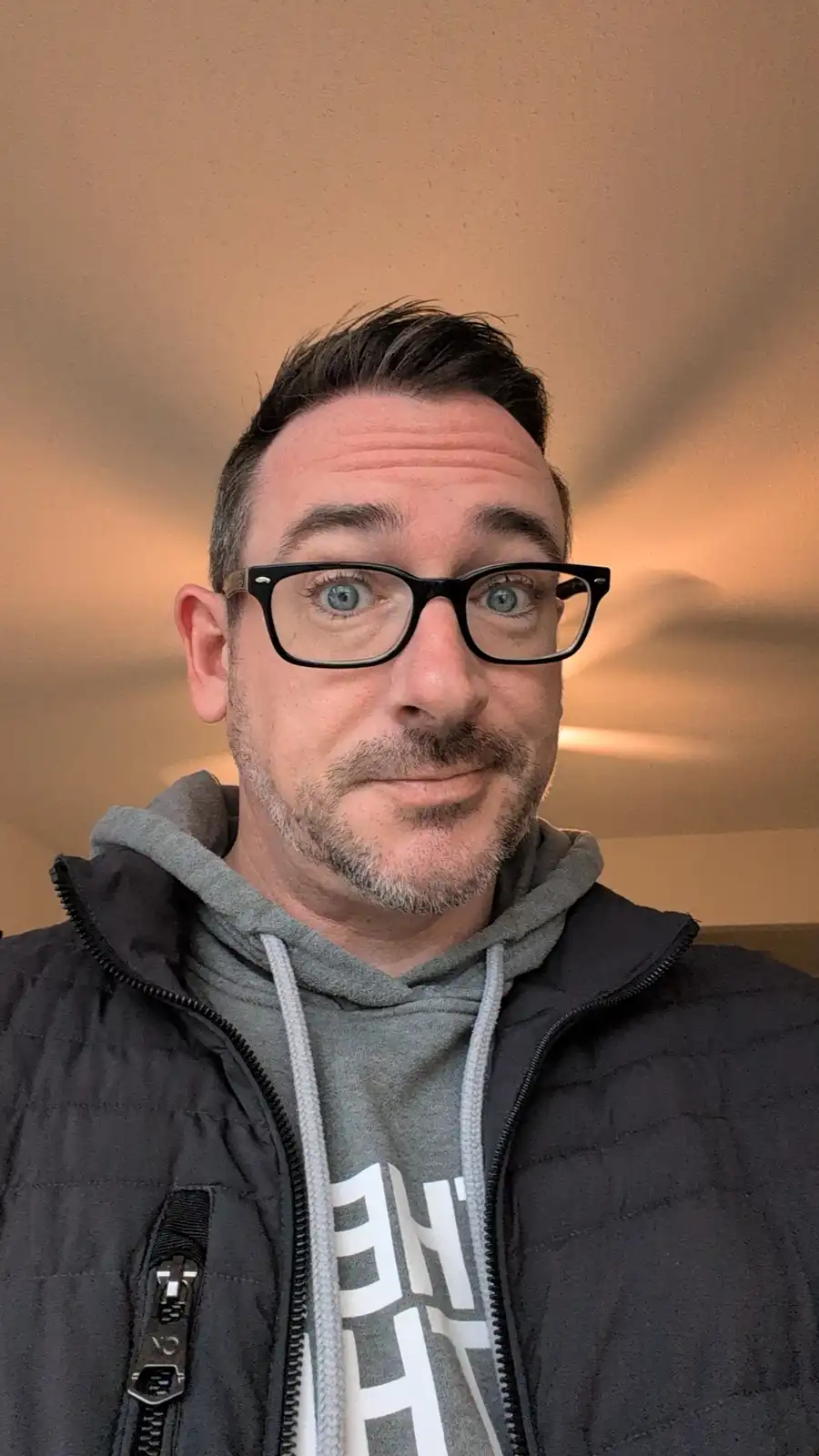 A man with blue eyes and black-rimmed glasses stares directly at the camera with a wide-eyed, slightly bewildered expression…