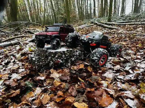 Two remote-controlled monster trucks, one red and one black with red rims, are positioned on a snowy forest stump surrounded…