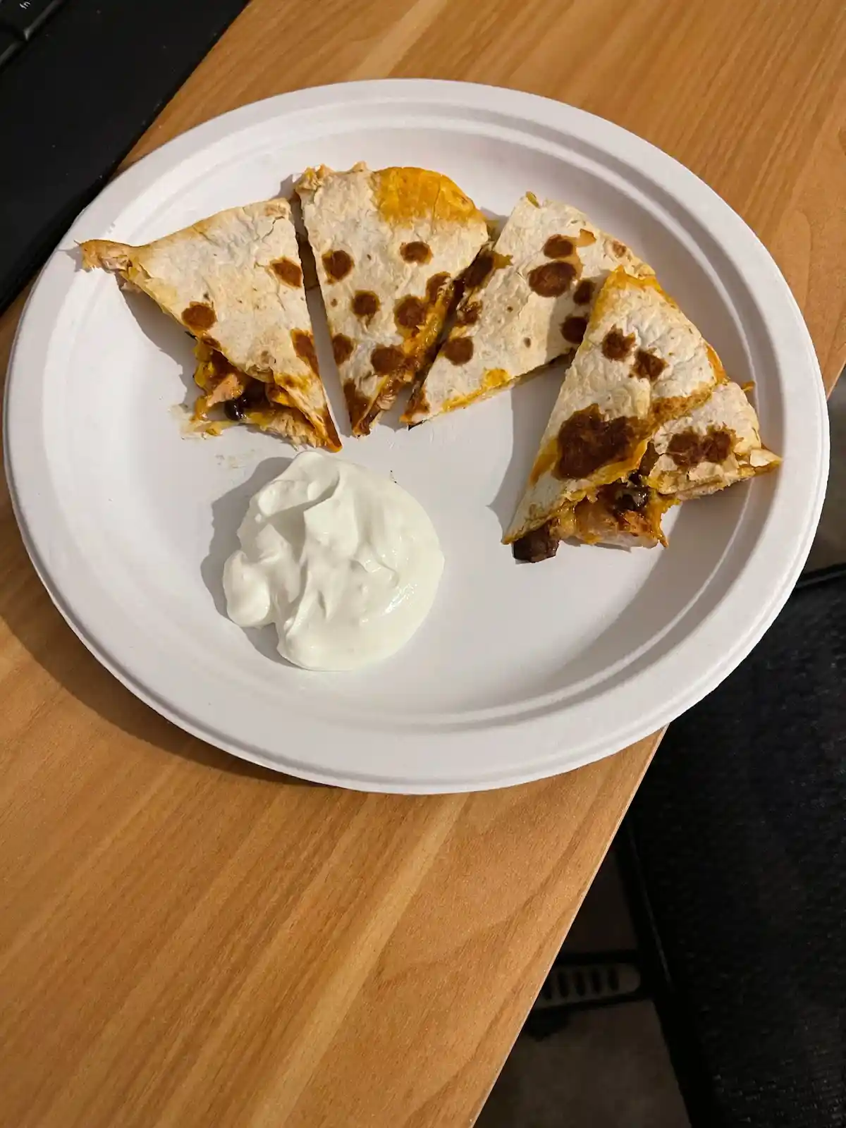 A white paper plate holds four triangular slices of a golden-brown quesadilla with melted cheese and dark fillings…