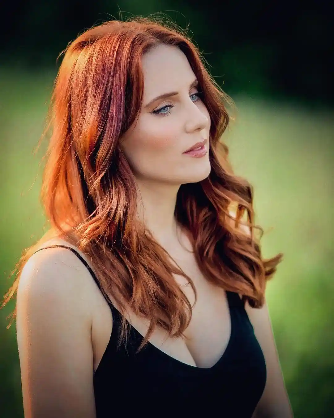 A woman with long, wavy, reddish-brown hair is shown in profile, looking off to the side. She wears a black tank top and has…