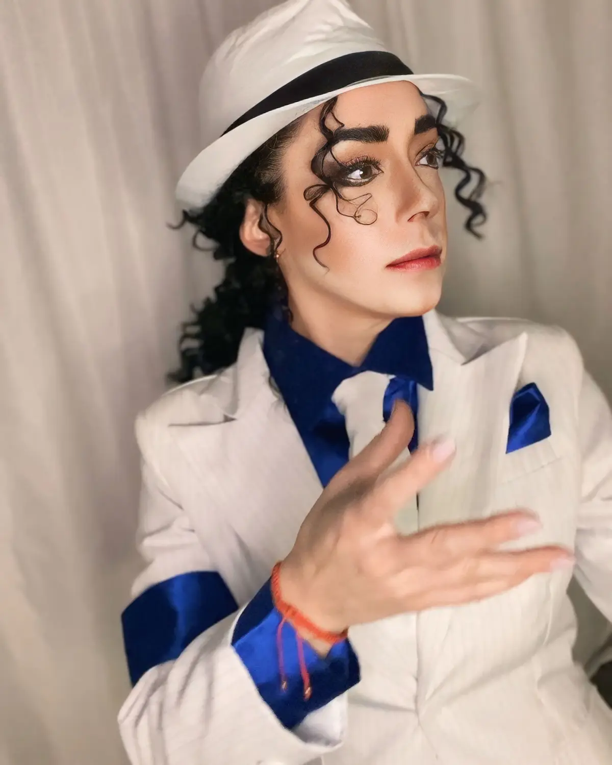 A person is dressed in a white pinstripe suit with blue satin accents, a white fedora with a black band, and has curly black…