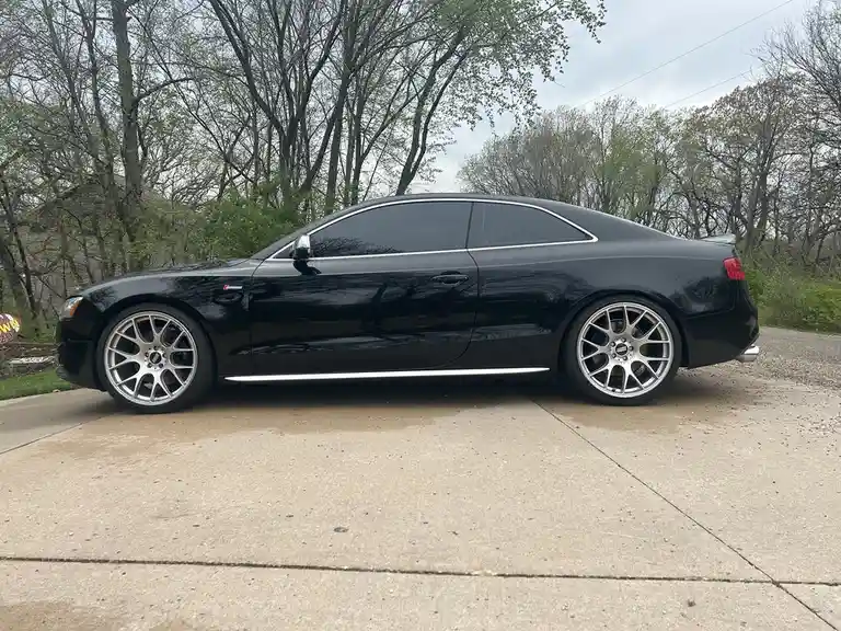 A black Audi S5 coupe with silver multi-spoke wheels is parked on a concrete driveway surrounded by trees. The car has a…
