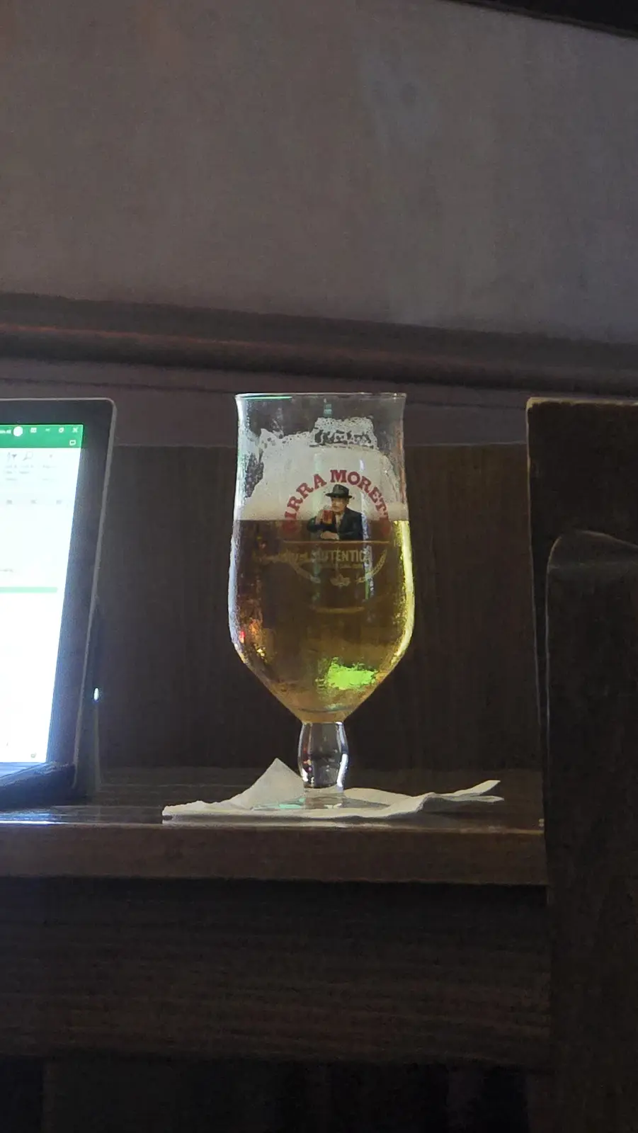 A half-full Birra Moretti beer glass with a frothy head sits on a napkin on a dark wooden table, next to a tablet displaying…